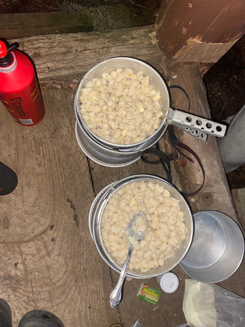 Two metal pots filled with mac on cheese on a wooden floor. There is a spoon in one of the pots.