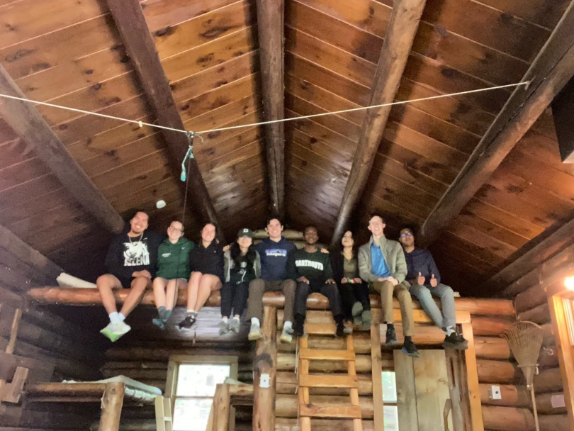 9 people sitting on a ledge overlooking the main part of the cabin. The ledge is connected to a ladder, and underneath the ledge is an alcove with beds.