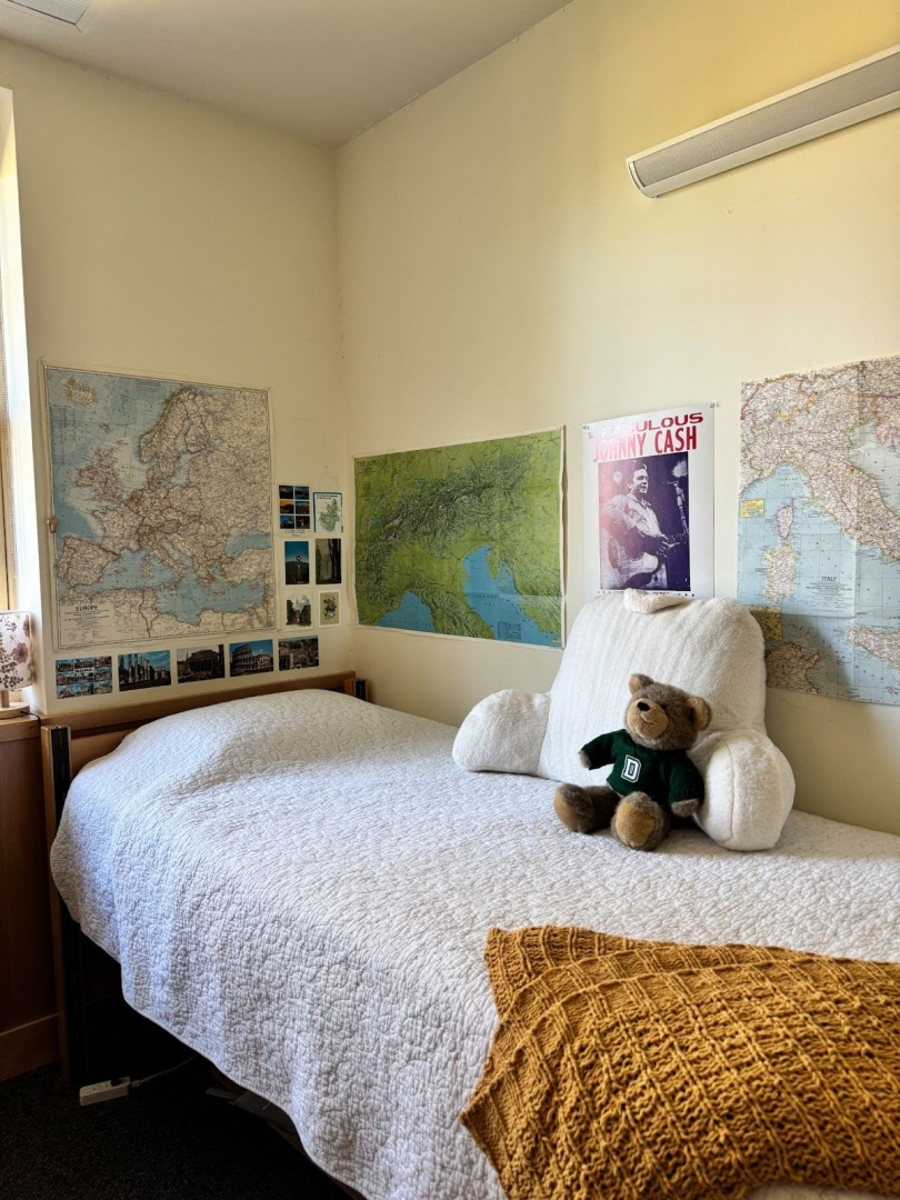 A photo of my bed, with the same stuffed bear animal and bedding. There are maps, postcards, and posters decorating the two walls behind the bed.