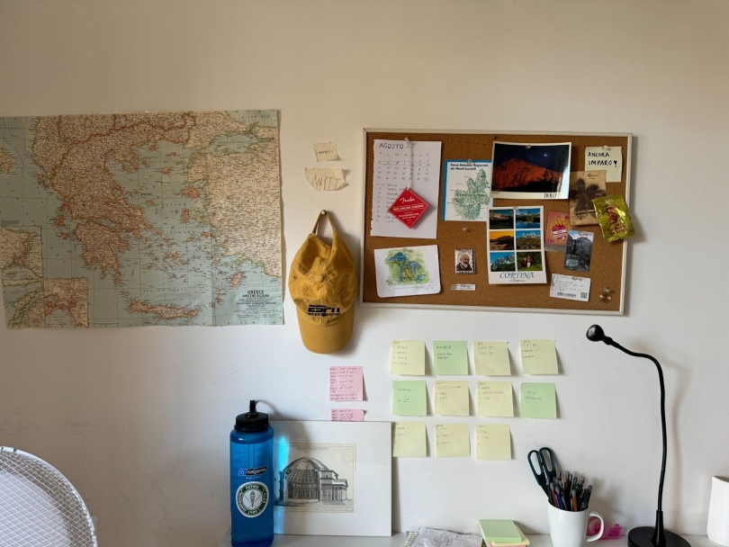 A photo of a bedroom wall, decorated with sticky notes, a map, and a cork board full of personal mementos. The edge of a desk is visible, with a water bottle, cup of pencils, and desk lamp on top of it.