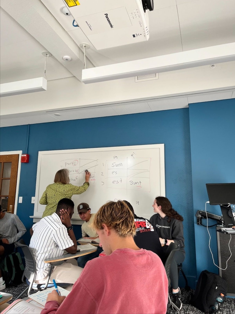 Students working in a small classroom while a professor writes on the whiteboard