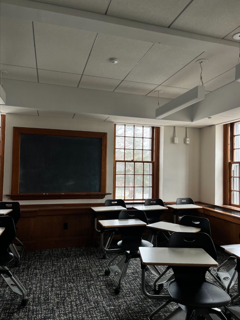An empty classroom with empty desks. The lights are turned off but light is coming in through the paned windows