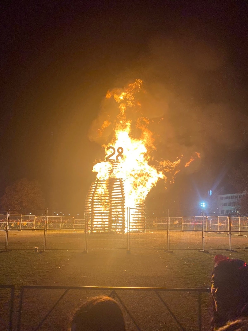 A bonfire made of overlapping wooden logs with a wooden "28" on the top of the bonfire. Bright orange flames encompass the structure and there is a fence around the bonfire.