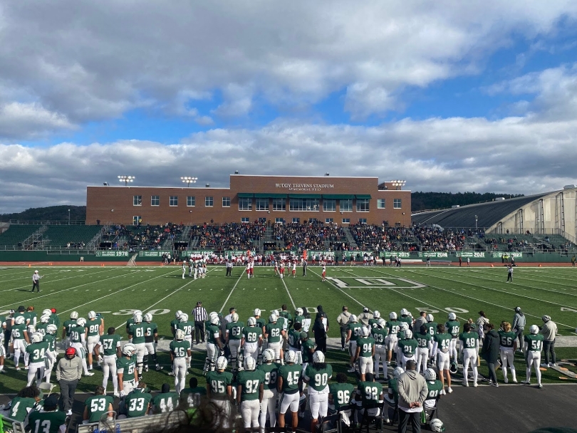 A large football field with a building in the background that says "Buddy Teevens Stadium at Memorial Field". In the foreground, there are football players wearing white helmets, green tops and white pants.