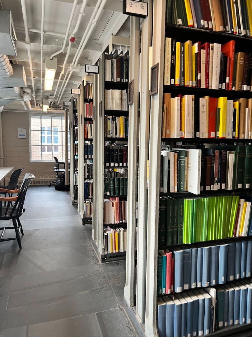 Bookshelves in the stacks full of colorful hardcover books. On the left side and in the distance, there are chairs at desks for students to study at. The chairs are empty.