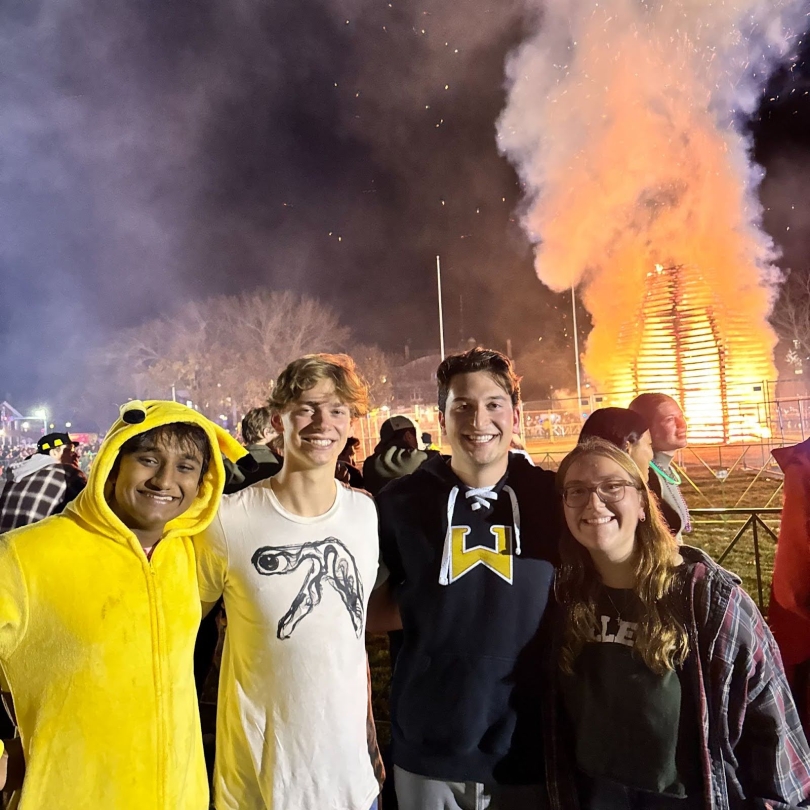 My friends Hrishik, Witt, Matt, and I standing in front of the Homecoming bonfire. It is dark outside and the fire is tall and bright in the background. Hrishik is wearing a Pokemon Halloween costume, and everyone is smiling at the camera.
