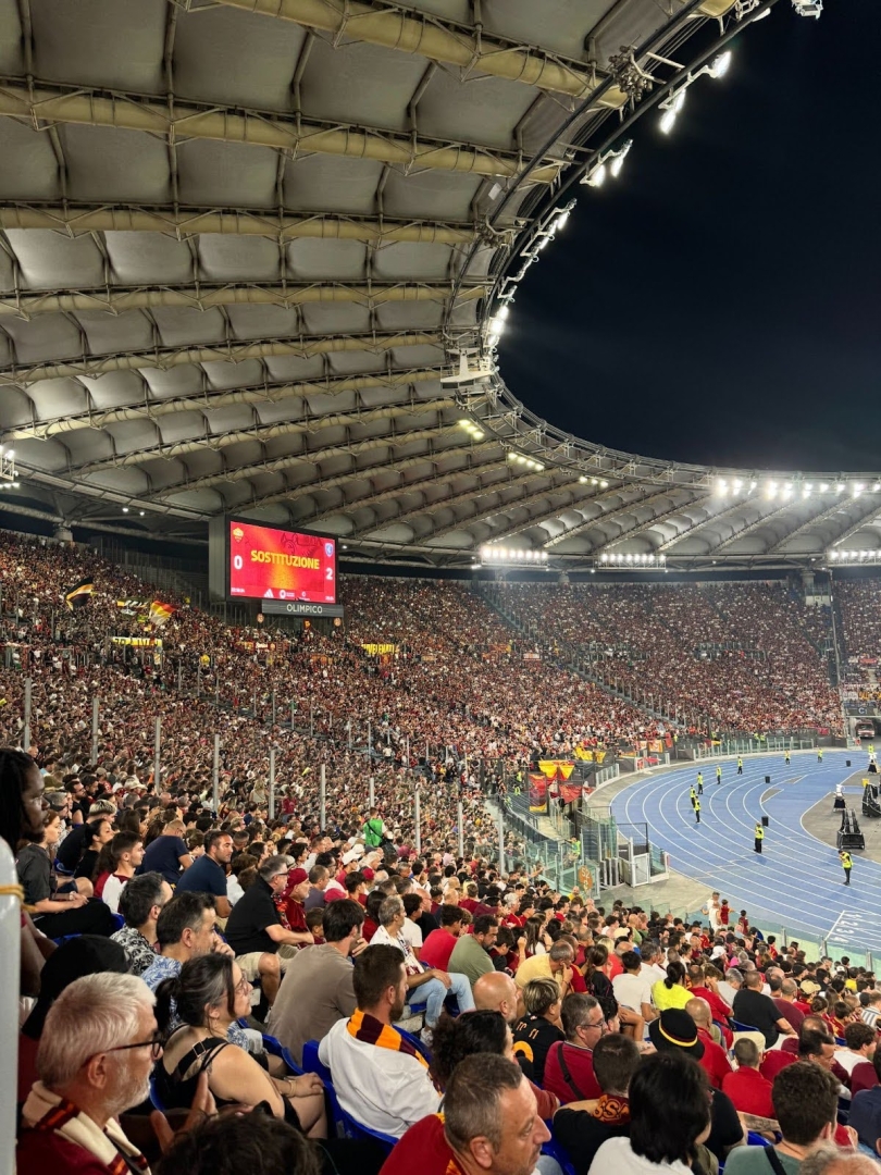 Thousands of people in the stands cheering on the AS Roma soccer club at their home opener. The stadium has an open roof, and you can see the night sky.