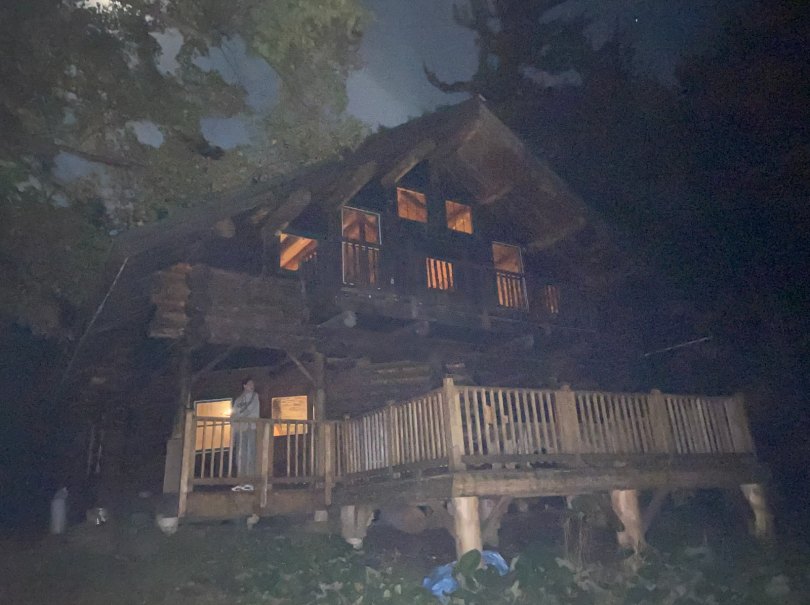  A large, two-story, wooden cabin with a balcony on the top and a large porch on the ground floor. The cabin has exposed beams and it is clear it was built from whole wooden logs. There are multiple windows on the cabin walls so the faint lights from insi