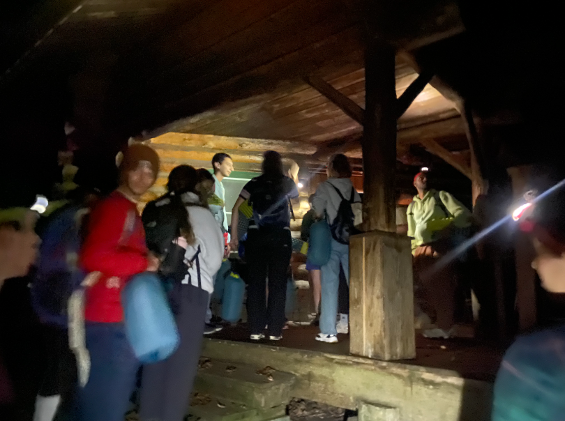 A group of people are standing on a porch entering a wooden cabin. Some people are hidden by the darkness of night while others are illuminated by headlamps and flashlights. The photo is a bit blurry but you can still make out the people standing together