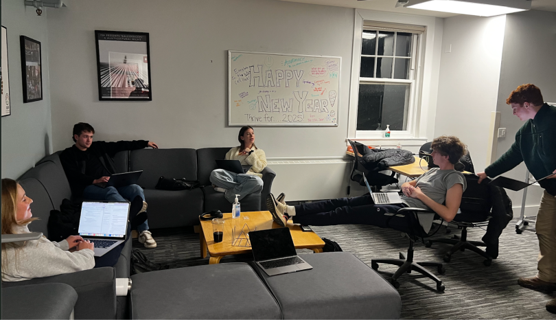 a group of people scattered around a living room with grey sofa and rolling chairs. With their laptops opened, they are in an intense meeting discussion.