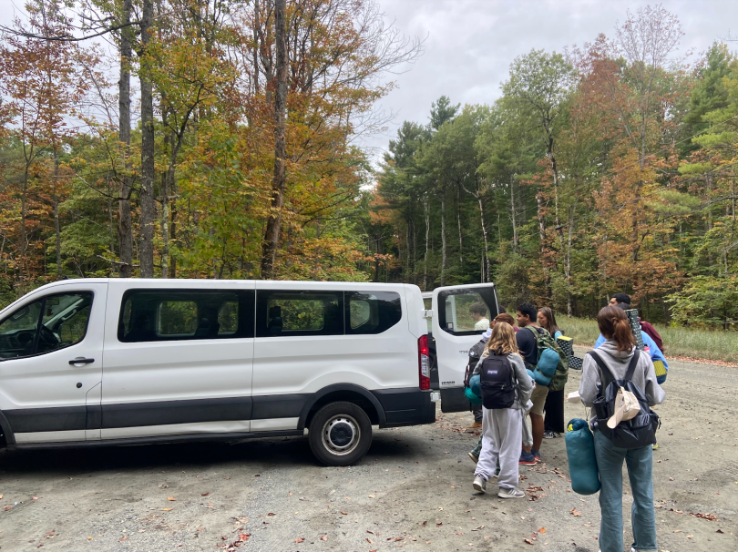 A white van is parked in a lot surrounded by trees. The leaves on the trees are changing colors. Some are green, some are orange, and others are red or yellow. The trunk of the van is open and a group of at least five people are gathered around the back. 