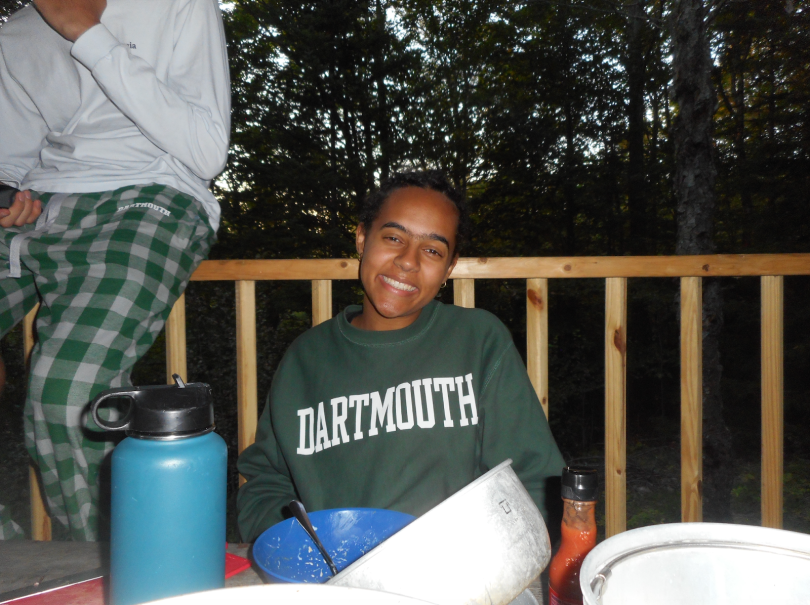 A girl, wearing a Dartmouth green sweatshirt, is smiling while sitting on the picnic table. In front of her on the table is a pile of used dishes. It is clear the girl has just finished cooking and eating dinner. Behind the girl is a wooden railing. 