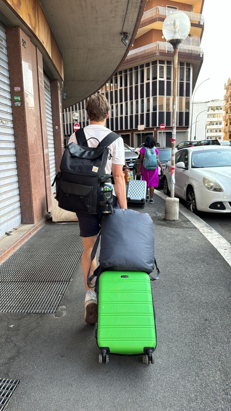 A boy wheeling a suitcase with a large bag on top of it while carrying a big backpack