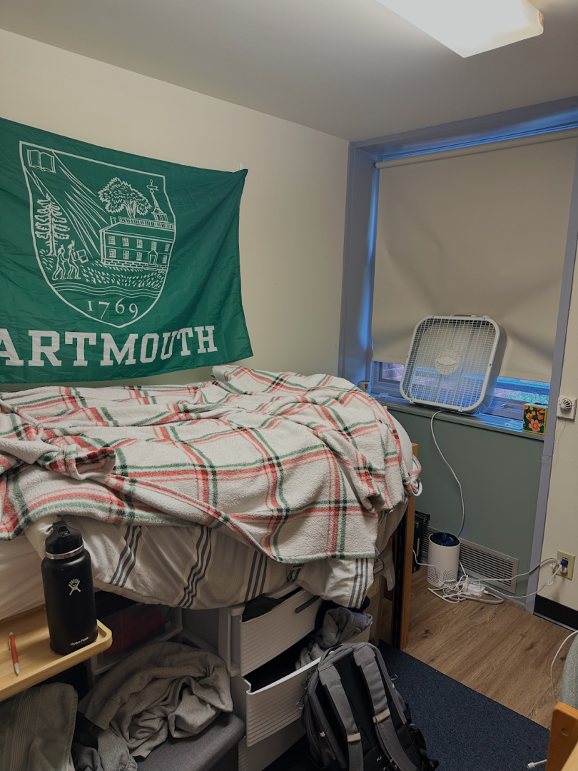 Dorm room with a bed covered in blankets, a Dartmouth flag on the wall, and wooden flooring.