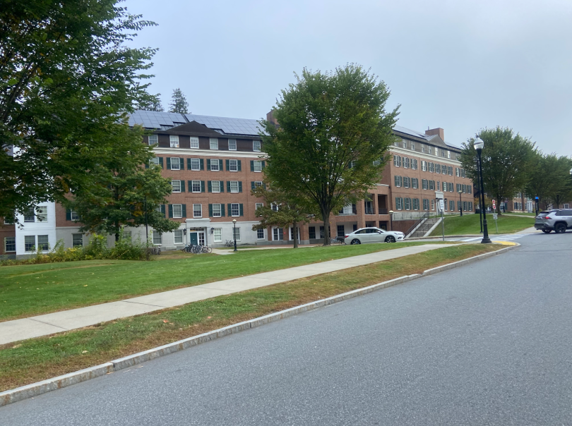 A long brick dorm building covers the pictures. In front of the building, there are four tall trees with really green leaves. There is also grass in front of the building and a sidewalk in front of the grass. The brick buildings are covered in windows. 