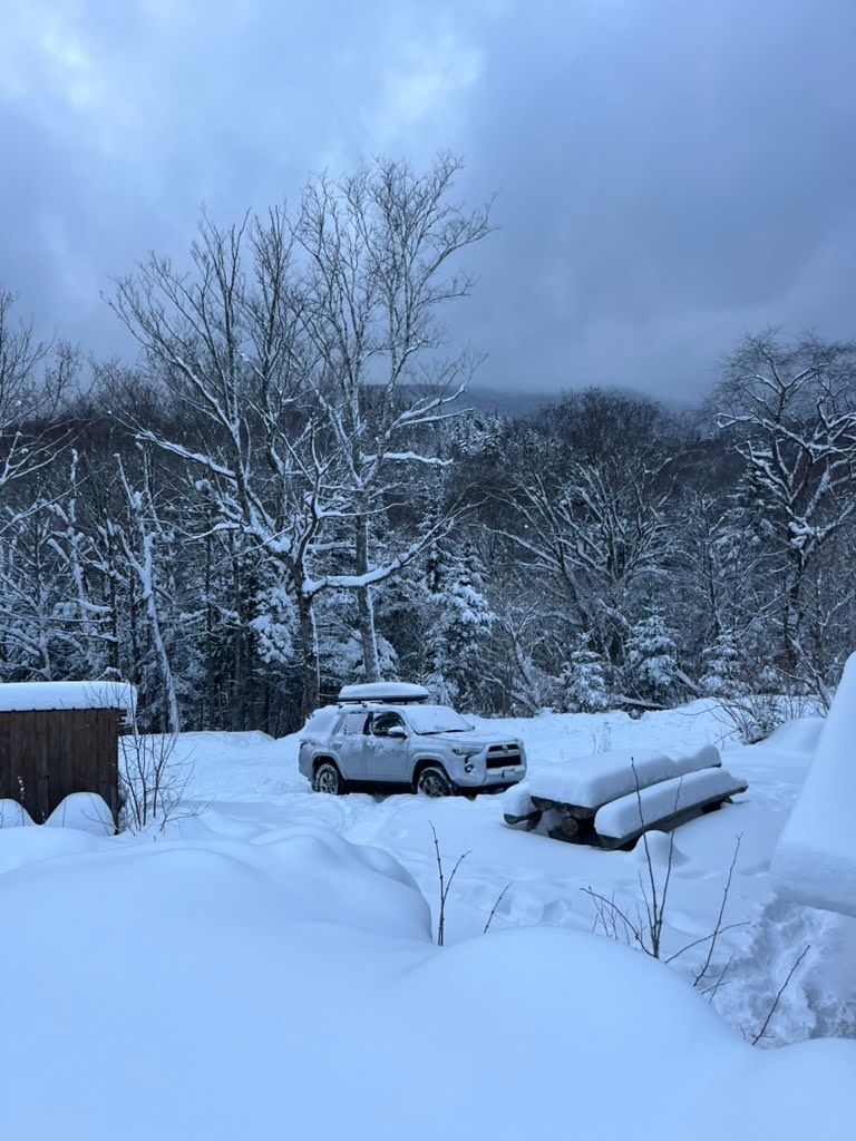 A photo looking at Mt. Moosilauke from the south side of the mountain. There is lots of snow. In the foreground, there is a shed, a picnic table, and a car. All are covered in snow.