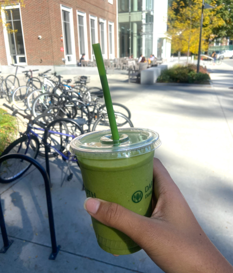  A green smoothie takes up most of the picture. Porturing from the top of the cup is a green straw. A hand is holding the drink, but you can only see the fingers and part of the palm. In the background, there is a brick building, gray concrete, and a bike