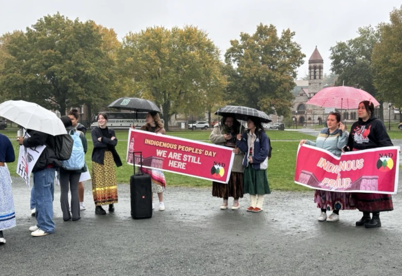 People stand under umbrellas holding "Indigenous Peoples' Day" posters