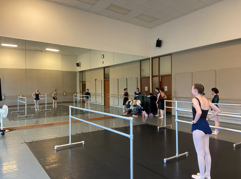 A large room with a wall of mirrors. In the center, there is a black rectangle on the floor. In the center of the room, there are three silver ballet barres. Ballet dancers are standing in the middle of the room, looking at the mirror.