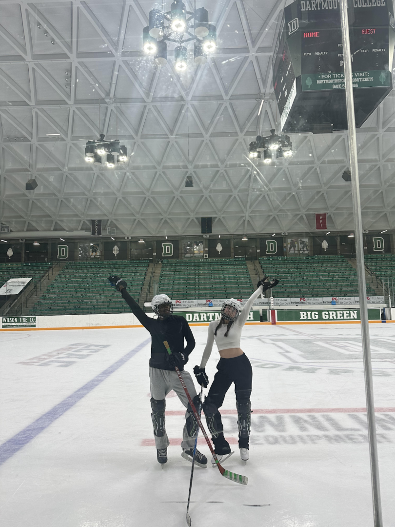  Two people standing in the center of the Dartmouth hockey ice rink, surrounded by the arena's vast, icy surface with the Dartmouth logo visible behind them. 