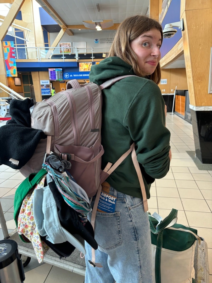  A girl in a green sweatshirt is looking at the camera. She is wearing a large backpack with many baseball caps attached to the outside of the bag.