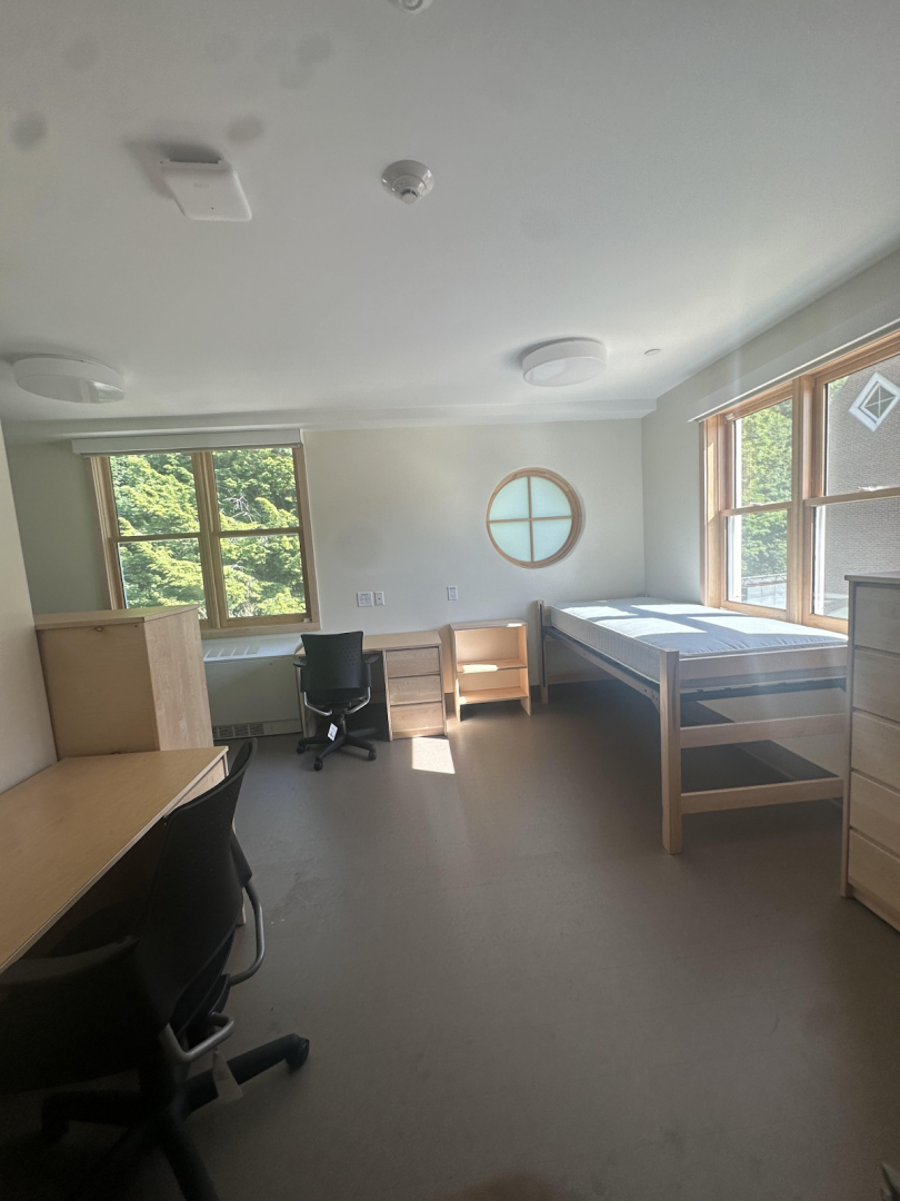 Vacant dorm room with two desks, two chairs, a bed, two dressers, and large windows letting in natural light.