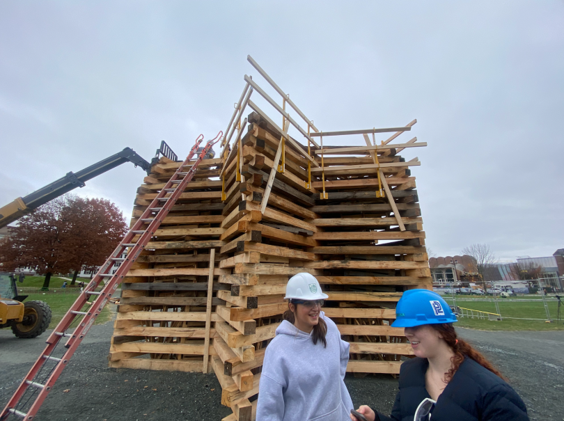A tall wooden structure is in the middle of the image. It is at least 1.5 stories tall. There is an orange ladder reaching the top that lies on the left, and there is a forklift to the left of the ladder. T