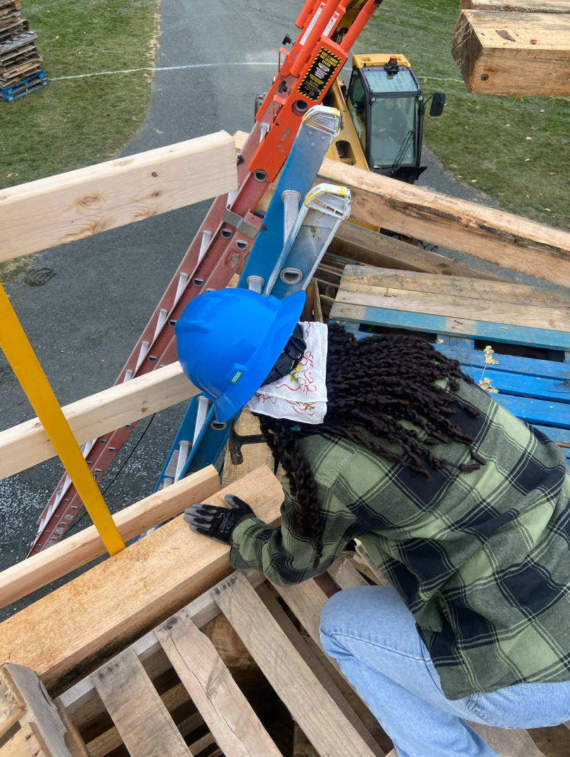 A girl wearing a blue hard hat and a black and green plaid shirt is crouched down on a wooden structure. She is holding a hammer and hitting a nail into a wooden beam.