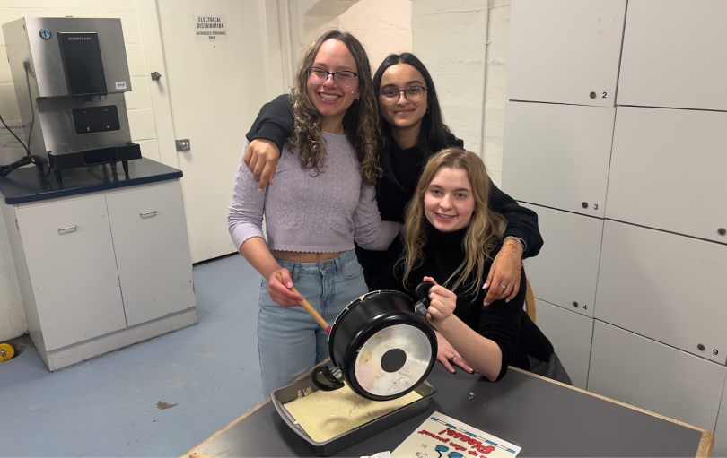 three girls smiling while holding a pot and spatula to spread the cake mix onto a pan