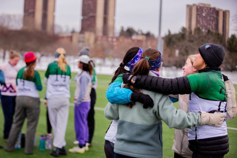 the camera focuses on dartmouth frisbee players huddling on the right while another group of dartmouth frisbee players on the left is slightly out of focus. They are standing on green grass. 