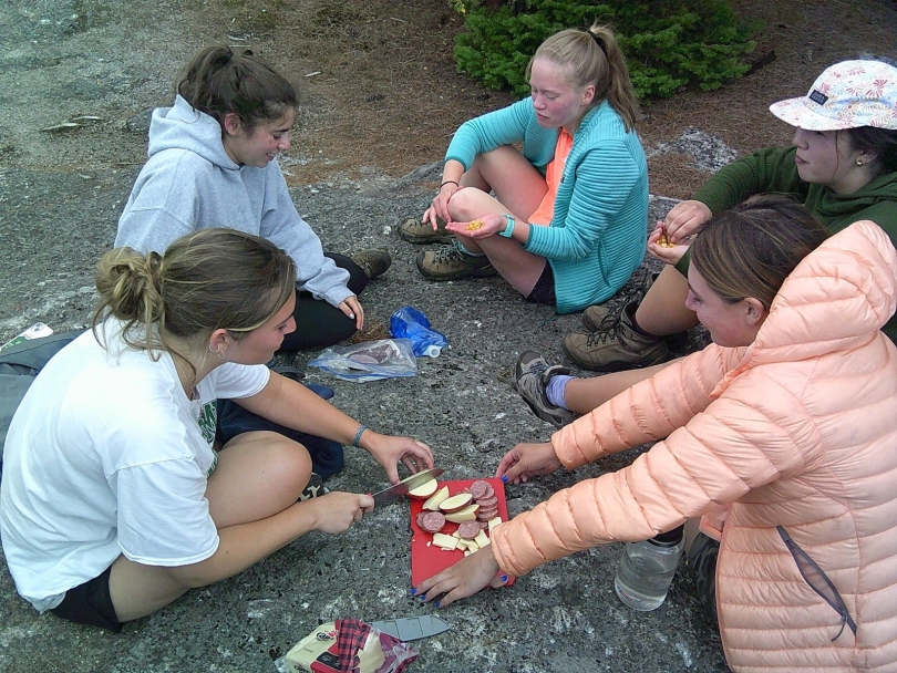 A group of humans sitting around granite rocks, at the summit of a trail. One person is slicing sausages, apples, and sharp cheese to compose a make-shift charcuterie board.
