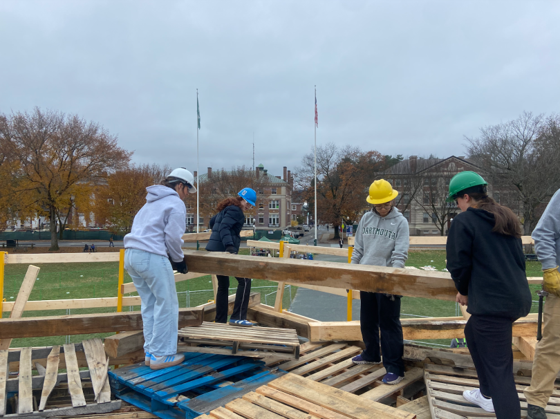 Four girls gather around a long wooden beam and hold it up. They are standing on wooden pallets. In the background, you can see barren trees, a cloudy sky, and two flagpoles.