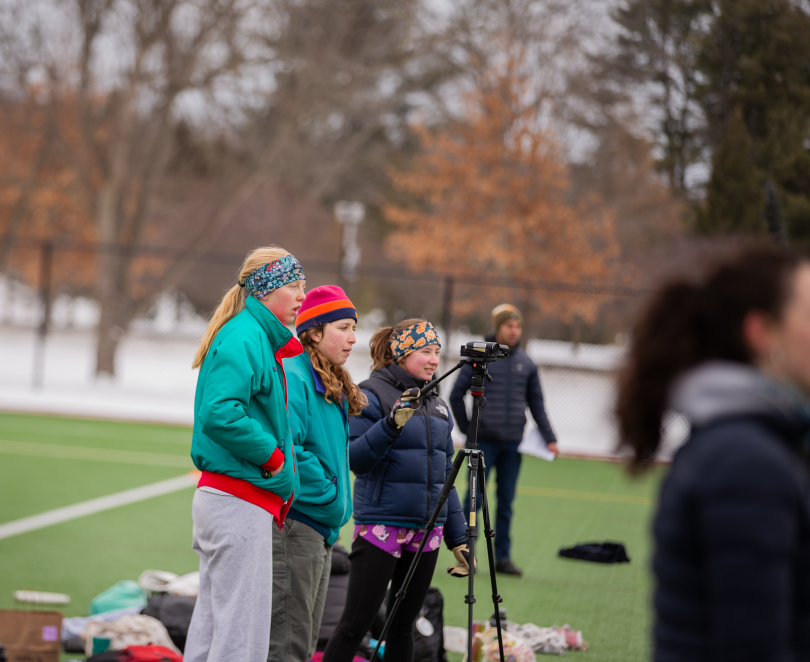 Three people stand in a line with a camera in front of them. They stand on green grass. 