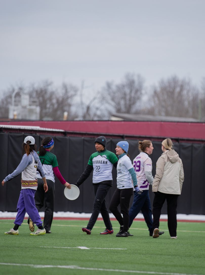 Six Dartmouth frisbee players walk in different directions. Two grasp a frisbee together. They are all standing on vibrant green grass. 