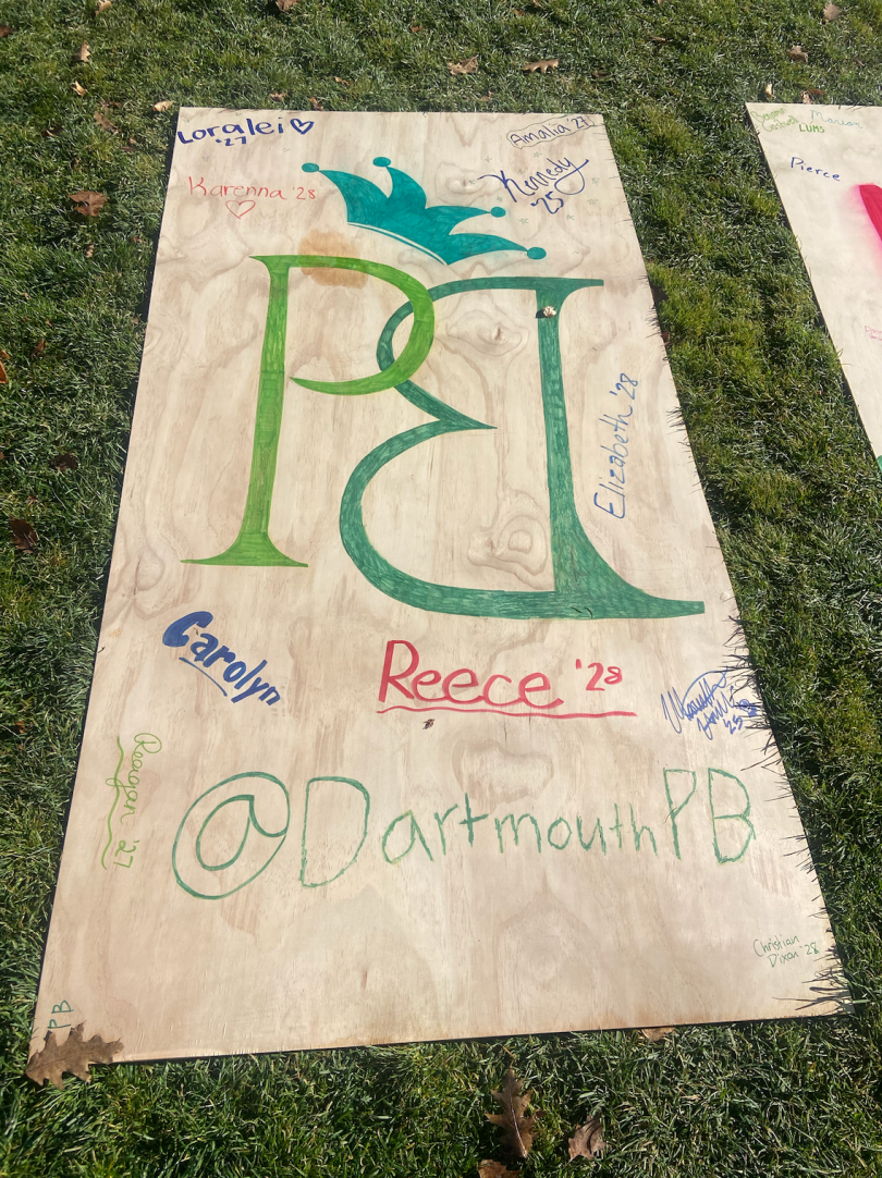 A wooden piece of plywood lies on green grass. A large "P" is drawn on the board in light green. Next to it, a large "B" is drawn backward in a darker green. On top of the "P," there is a teal crown. 