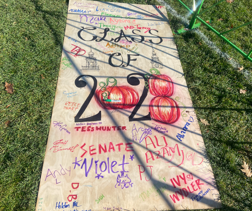 A wooden piece of plywood lies on green grass. Three large words in the center read "Class of 2028." The "0" and "8" in the year are made of orange pumpkins. Various people have signed their names using different-colored markers surrounding the text.