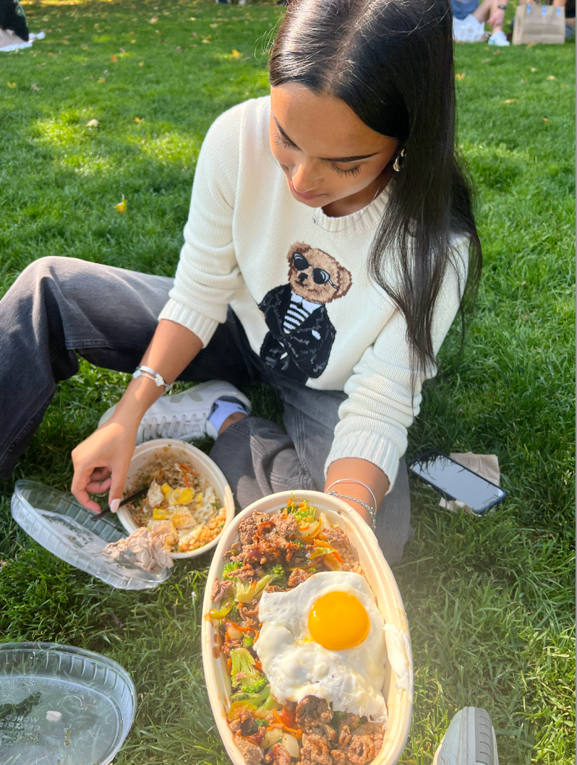 A girl sitting by a plain of grass, holding a bowl of stir fry with a sunny-side up egg. 