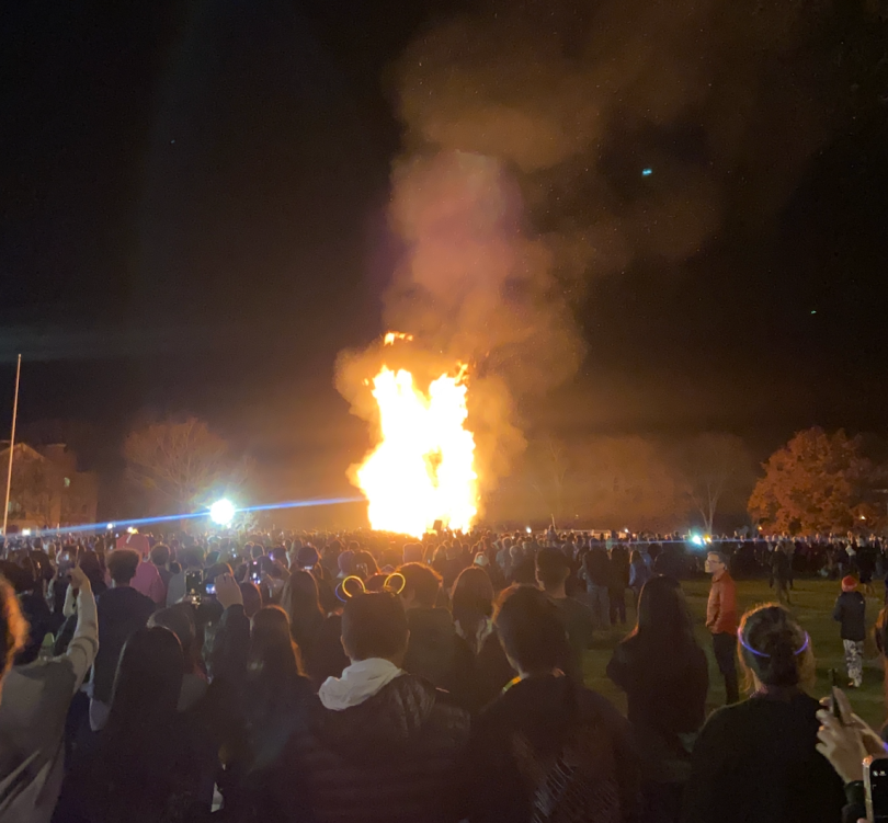 A tall burning structure is in the middle of the image. Billows of smoke rise from the fire, and large crowds of people surround it. It is night so the sky is very dark.