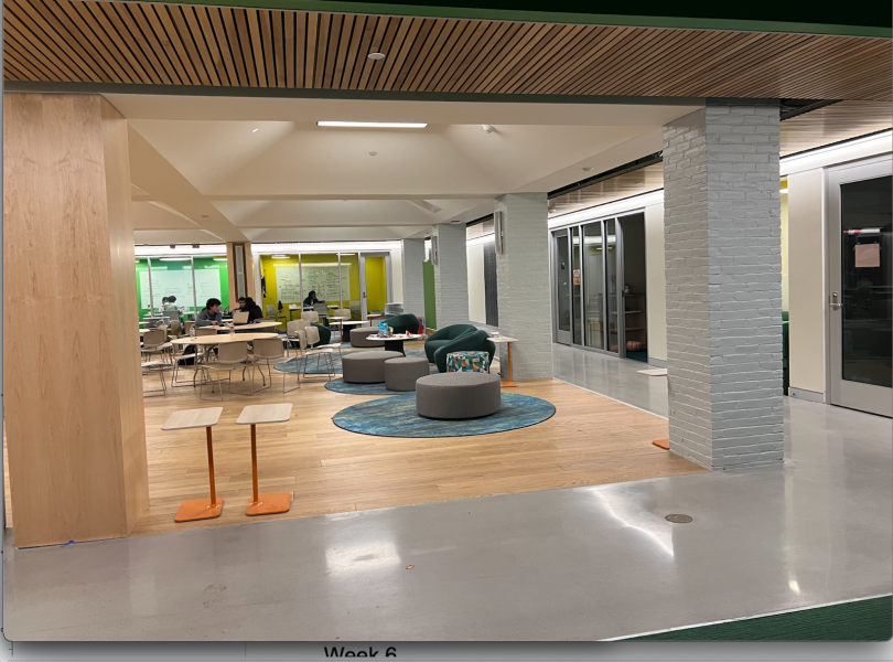 An open space with stools on carpet, wooden desks, and green sofas. Surrounding the space are glass walls leading into study rooms