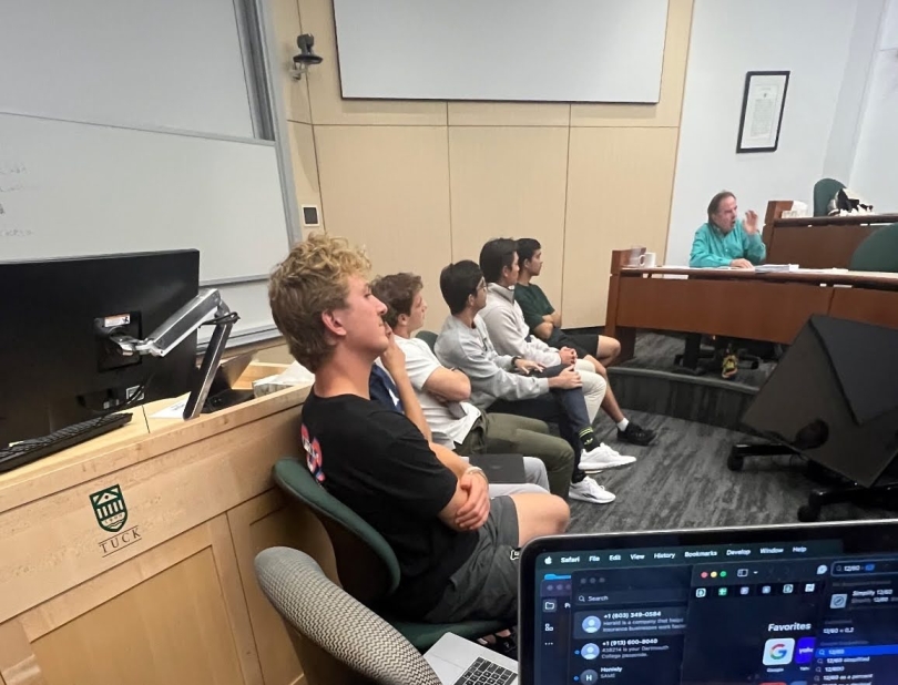 A presentation floor seated by 7 people all facing towards the man dressed in cyan giving feedback to their presentation.