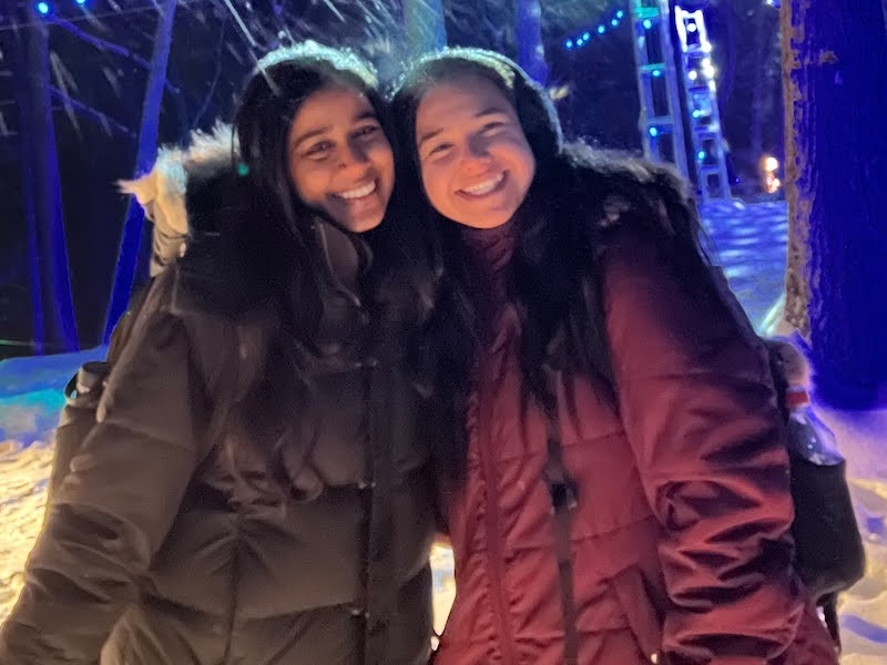 An image of two students in winter jackets in a forest with snow on the ground.