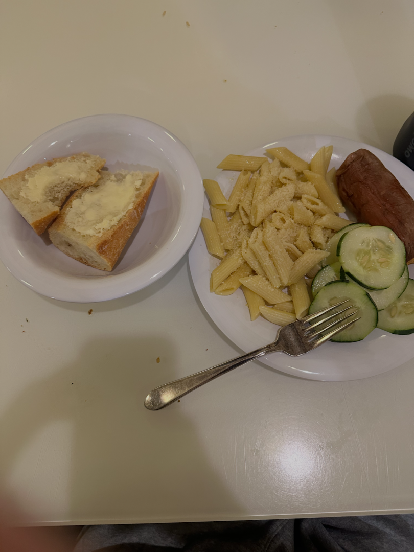A plate with noodles, a potato, and cucumbers on it and a bowl with buttered bread.