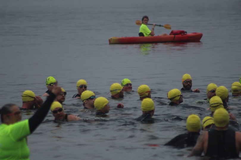 Triathletes in yellow swim caps stand neck deep in water waiting to swim.