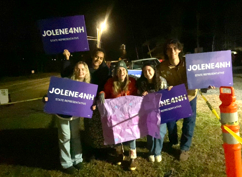 A group of five people stand together holding purple yard signs that read "JOLENE4NH." It is dark outside, and they are standing on a grassy patch. 