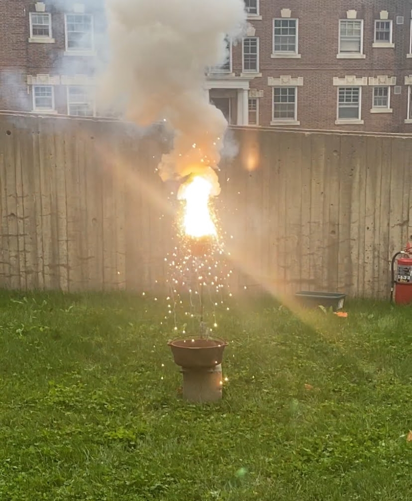 A clay pot emitting fire, smoke and sparks. The pot is sitting on a grass lawn and there is a wall in the background.