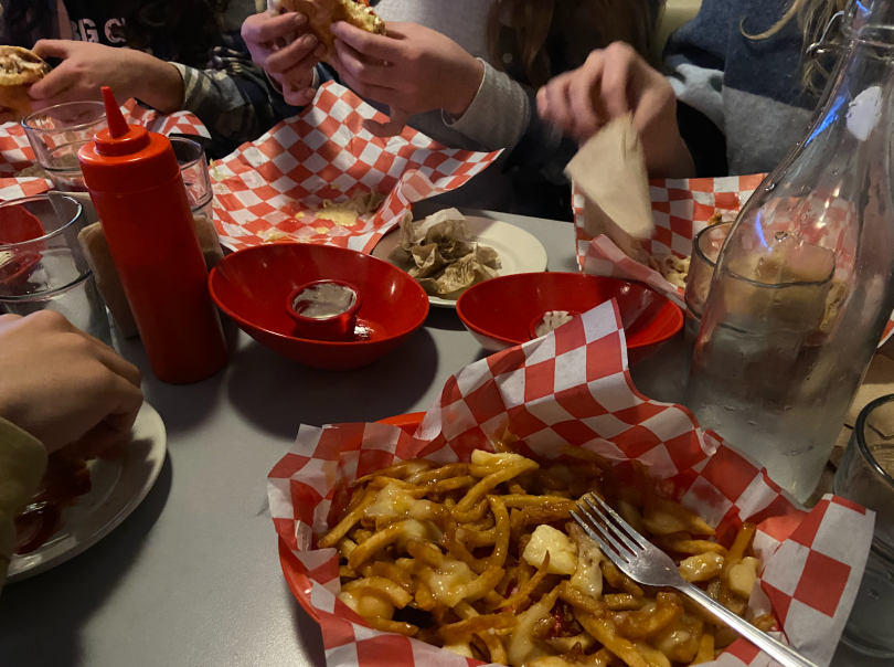 Fried and cheese curds are in a diner basket that is lined with red and white checkerboard paper. The diner table is full of similar foods. There is a ketchup bottle, a red bowl for fried pickles, a glass water canteen, and much more! 