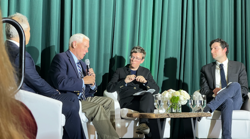 Four people sit on a stage; each is dressed in business attire. The second person to the left, Mike Pence, holds a microphone. Behind them is a green curtain, and in front of them, there is a table with white flowers.