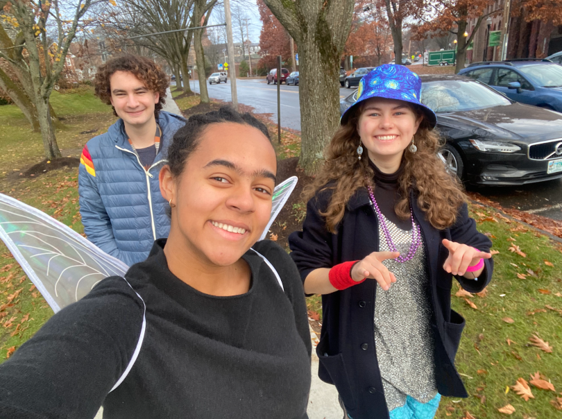 A girl in a black sweater and fairy wings is next to another girl wearing a sequin silver dress, a blue bucket hat, and a black coat. Behind them, there is a man in a blue jacket. The photo is a selfie.