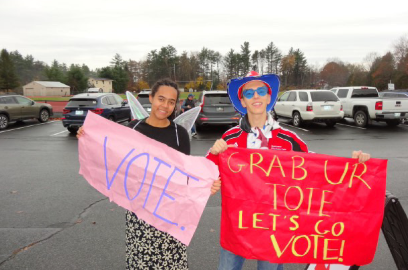 A girl wearing fairy wings holds a pink sign that reads "VOTE." while a man, in glasses and a cowboy hat, holds one that reads "grab ur tote let's go vote!"