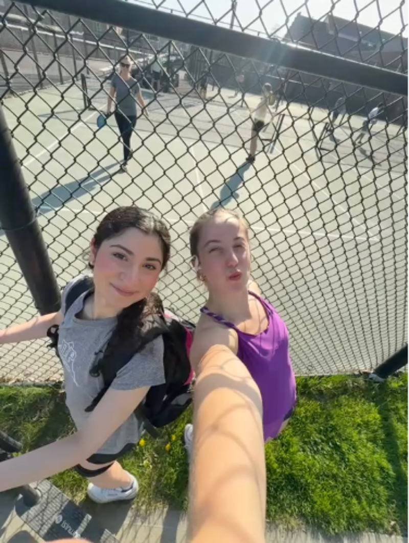 two people taking a 0.5x selfie with people playing pickleball
